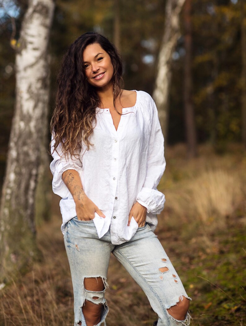 May include: A woman with long, wavy brown hair is wearing a white linen shirt and light blue jeans with distressed details. She is standing in a forest setting with trees in the background.