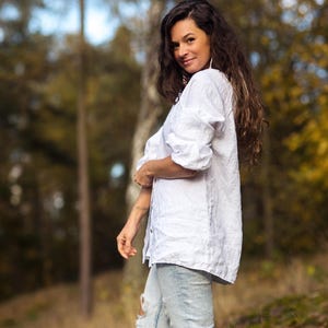 May include: A woman wearing a white linen shirt and light blue jeans with distressed details. She is standing in a forest with trees in the background.