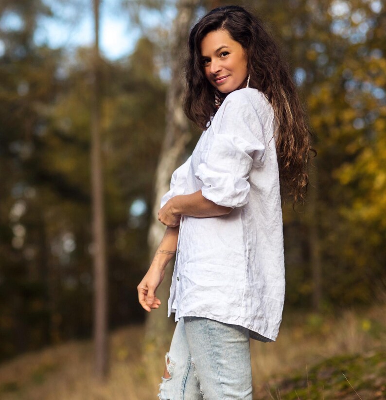 May include: A woman wearing a white linen shirt and light blue jeans with distressed details. She is standing in a forest with trees in the background.