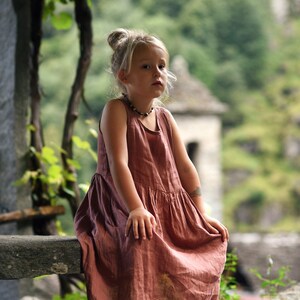 May include: A young child wearing a sleeveless, dusty rose-colored dress sits on a stone ledge. The dress has a gathered skirt and a simple neckline. The child has blonde hair pulled up in a bun and is wearing a beaded necklace.