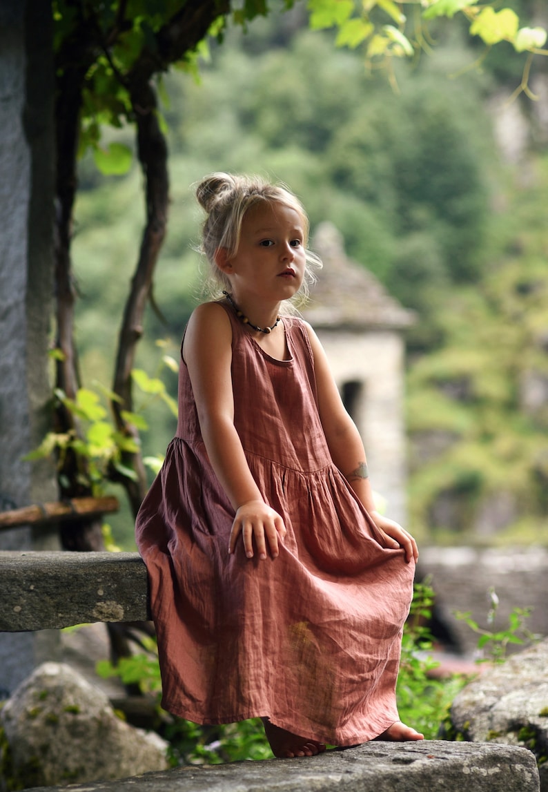 May include: A young child wearing a sleeveless, dusty rose-colored dress sits on a stone ledge. The dress has a gathered skirt and a simple neckline. The child has blonde hair pulled up in a bun and is wearing a beaded necklace.