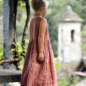 May include: A sleeveless, dusty rose-colored dress with a gathered waist. The dress is worn by a young person with their hair in a bun. The background features a stone structure and lush greenery, suggesting an outdoor setting.