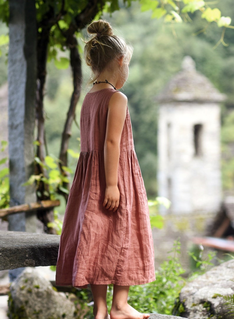 May include: A sleeveless, dusty rose-colored dress with a gathered waist. The dress is worn by a young person with their hair in a bun. The background features a stone structure and lush greenery, suggesting an outdoor setting.