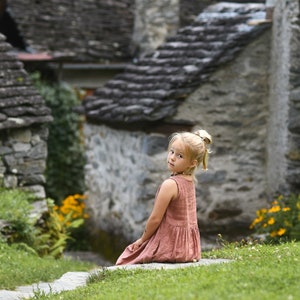May include: A young girl with blonde hair in a ponytail sits on a stone step, looking back at the camera. She wears a sleeveless, dusty rose dress. The background features stone buildings and green grass with yellow flowers.