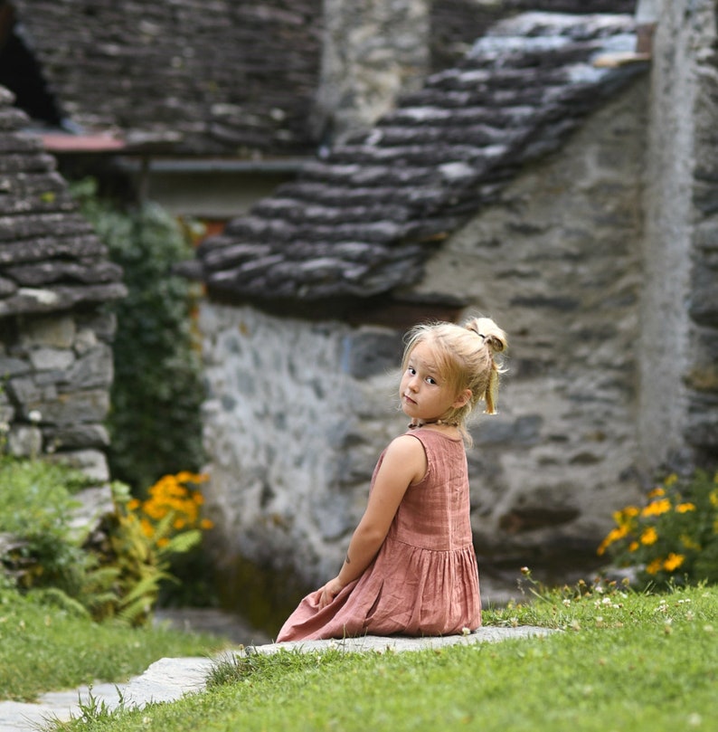 May include: A young girl with blonde hair in a ponytail sits on a stone step, looking back at the camera. She wears a sleeveless, dusty rose dress. The background features stone buildings and green grass with yellow flowers.