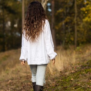 May include: A woman with long, curly brown hair walks through a forest wearing a white linen shirt, light blue jeans, and black boots.