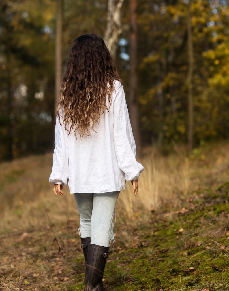 May include: A woman with long, curly brown hair walks through a forest wearing a white linen shirt, light blue jeans, and black boots.