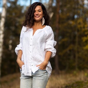 May include: A woman wearing a white linen shirt with a button-down front and a pair of light blue distressed jeans. She is standing in a forest setting with trees in the background.