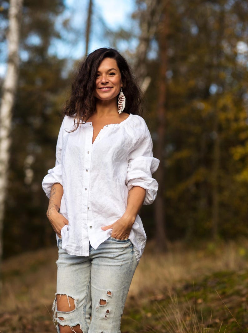 May include: A woman wearing a white linen shirt with a button-down front and a pair of light blue distressed jeans. She is standing in a forest setting with trees in the background.