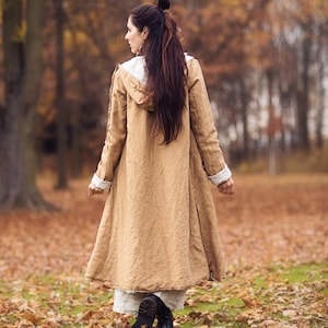 Può includere: Un lungo cappotto beige con cappuccio e finiture bianche è indossato all'aperto. Il cappotto è lungo fino al ginocchio e ha un design fluido. La persona si allontana dalla fotocamera in un parco con foglie cadute a terra.