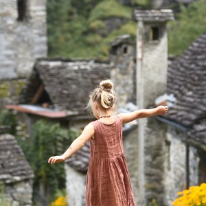 May include: A young person in a rust-colored sleeveless dress stands on a stone wall with arms outstretched. The background features a stone village with slate roofs and lush greenery, and yellow flowers.