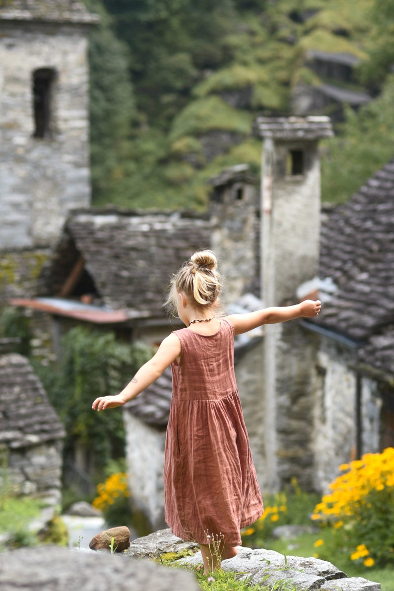 May include: A young person in a rust-colored sleeveless dress stands on a stone wall with arms outstretched. The background features a stone village with slate roofs and lush greenery, and yellow flowers.