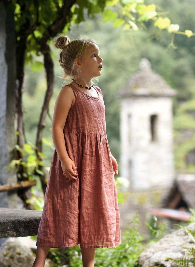 May include: A young girl wearing a sleeveless, dusty rose-colored dress. The dress has a gathered waist and falls to mid-calf. She is looking upwards with a thoughtful expression. A small beaded necklace adorns her neck. The background features a blurred stone building and lush greenery.