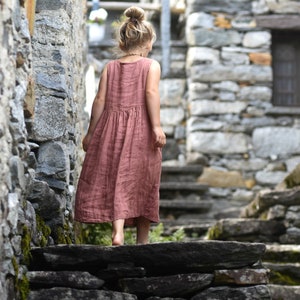 May include: A young person wearing a sleeveless, dusty rose-colored dress walks up stone steps in a rustic, stone-walled alley. The dress has a gathered waist and falls to mid-calf. The person has blonde hair in a bun.