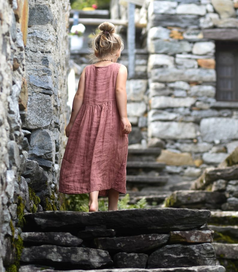 May include: A young person wearing a sleeveless, dusty rose-colored dress walks up stone steps in a rustic, stone-walled alley. The dress has a gathered waist and falls to mid-calf. The person has blonde hair in a bun.