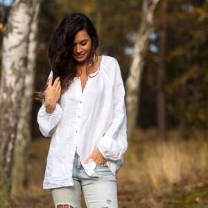May include: A woman wearing a white linen shirt with a relaxed fit and distressed blue jeans. She is standing in a forest setting with birch trees in the background.
