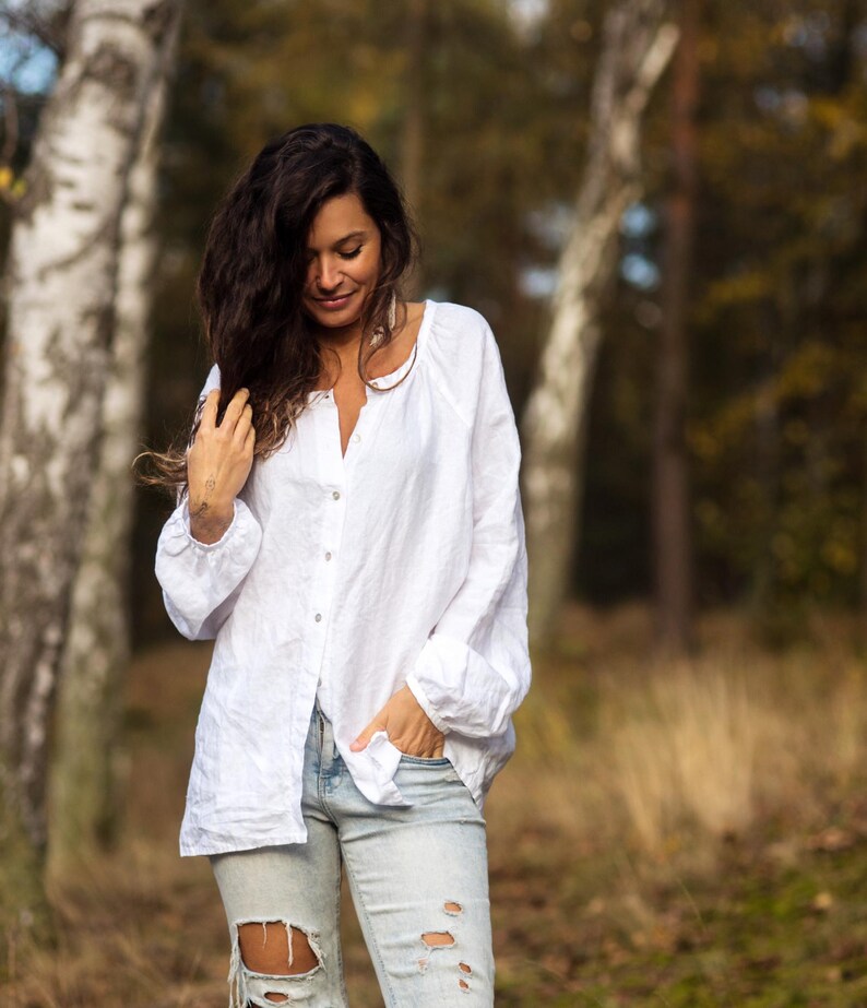 May include: A woman wearing a white linen shirt with a relaxed fit and distressed blue jeans. She is standing in a forest setting with birch trees in the background.