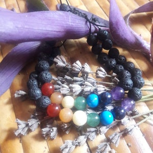 Op de afbeelding: Een armband met edelstenen in chakra-kleuren en zwarte lavabeads. De armband wordt gepresenteerd met gedroogde lavendel takjes en paarse bladeren op een bamboe oppervlak. De edelstenen zijn rood, oranje, geel, groen, blauw en paars.