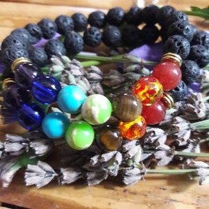 May include: Two beaded bracelets with a variety of colorful stones and black lava beads. The bracelets are arranged on a bed of dried lavender. The colorful beads include blue, turquoise, green, brown, orange, and red.