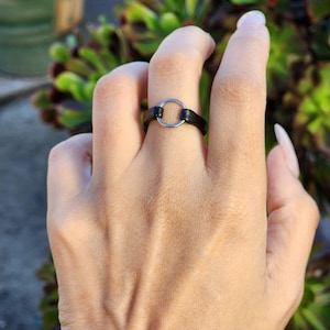 Puede incluir: Un anillo de cuero negro con un diseño de círculo plateado. El anillo se lleva en un dedo, con un fondo borroso de plantas verdes. El anillo tiene un diseño simple y minimalista.