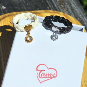 Könnte beinhalten: Zwei geflochtene Ringe, einer cremefarben und einer schwarz, jeweils mit einem Anhänger und einem klaren Edelstein. Die Ringe liegen auf einer weißen Schachtel mit einem roten Herzlogo mit der Aufschrift "Tame".