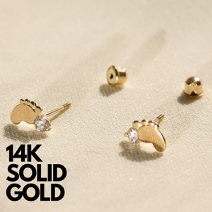 May include: A pair of 14K solid gold baby footprint earrings with a single clear cubic zirconia stone on each earring.