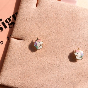 May include: A pair of gold unicorn stud earrings on a pink velvet background. The unicorn is white with pink, blue, and purple accents.