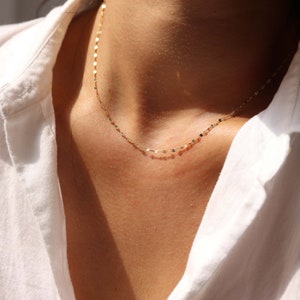 May include: A delicate gold chain necklace with a delicate, flat, linked chain. The chain is worn by a person with a white shirt.