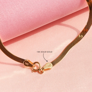 May include: A close-up of a 14k solid gold snake chain necklace. The chain is made of a thin, flat, and flexible metal. The clasp is a lobster claw clasp. The chain is on a pink fabric background.