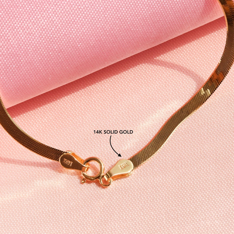 May include: A close-up of a 14k solid gold snake chain necklace. The chain is made of a thin, flat, and flexible metal. The clasp is a lobster claw clasp. The chain is on a pink fabric background.