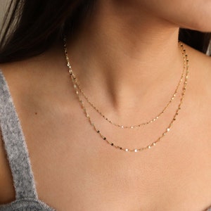 May include: A gold chain necklace with a delicate, layered design. The chain is made of small, flat, oval-shaped links.