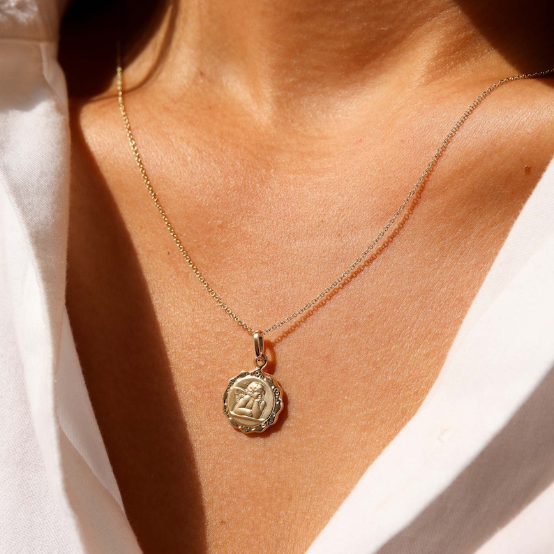 May include: A gold chain necklace with a small, round pendant featuring a detailed image of an angel. The pendant has a textured, scalloped edge.