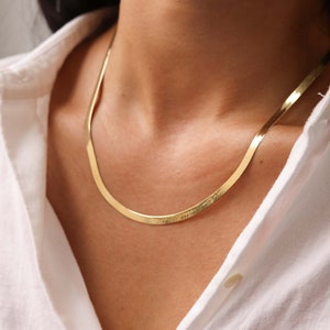 May include: A gold chain necklace with a flat, snake-like design. The chain is worn by a person with a white shirt.
