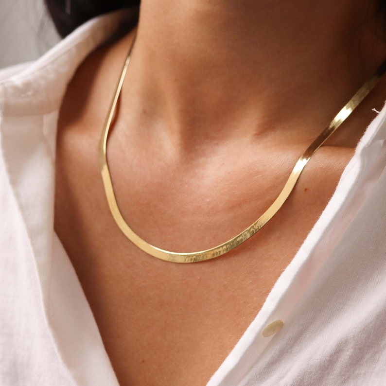 May include: A gold chain necklace with a flat, snake-like design. The chain is worn by a person with a white shirt.