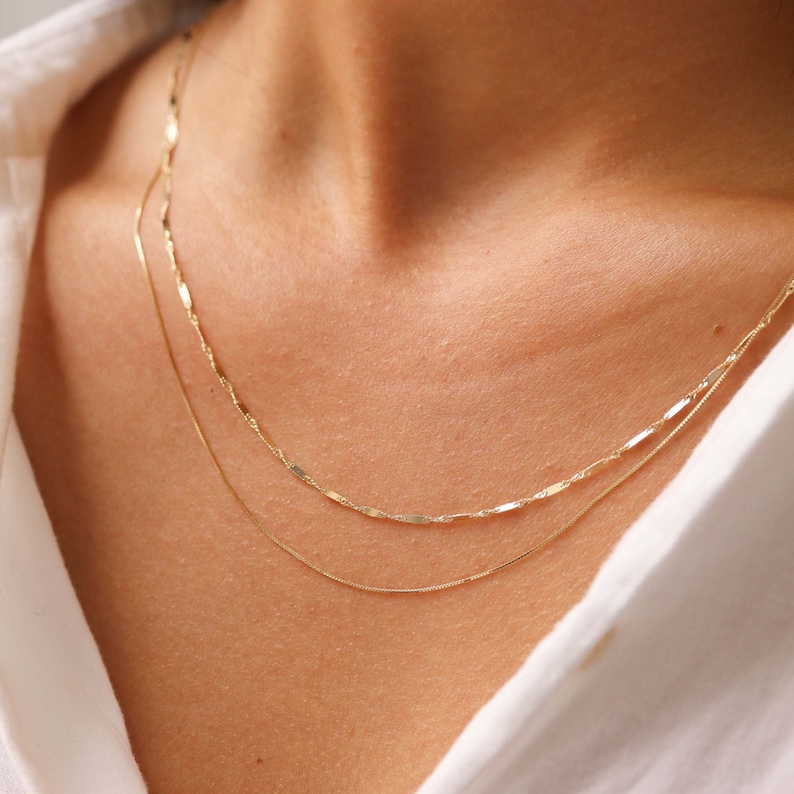 Pu&ograve; includere: Due collane d'oro sovrapposte sul collo di una persona. La collana superiore ha una catena delicata e piatta con un anello leggermente ovale. La collana inferiore &egrave; una catena rotonda pi&ugrave; sottile.