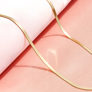 May include: A gold chain necklace with a flat, thin, and shiny design. The chain is lying on a pink surface.