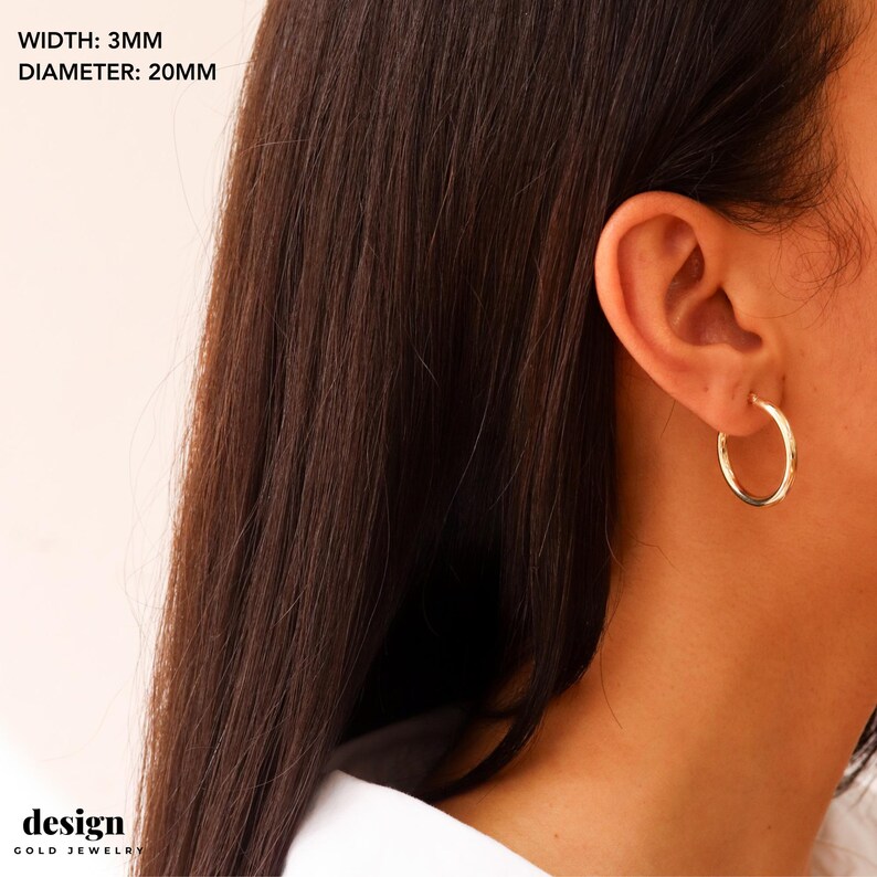 May include: Gold hoop earrings with a 20 mm diameter and 3 mm width. The earrings are shown on a person's ear.