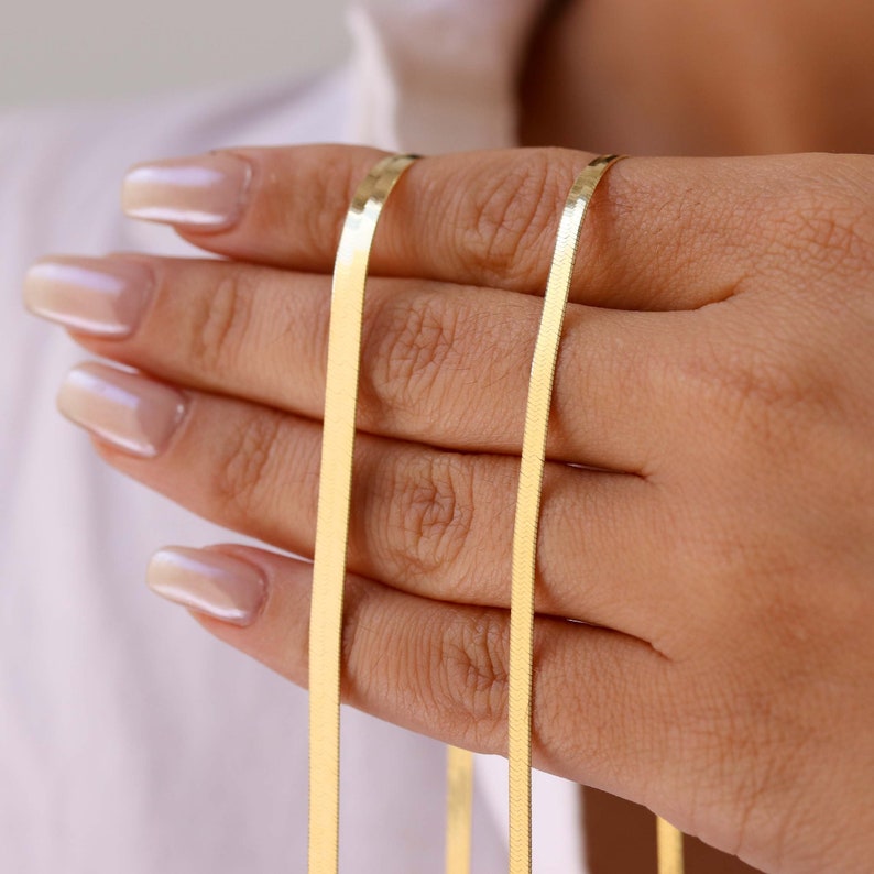 May include: Two gold-colored snake chains, one thicker than the other, are held in a hand. The chains are smooth and shiny.
