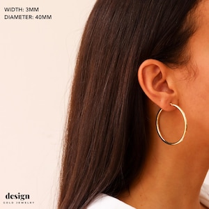 May include: Gold hoop earrings with a 40mm diameter and 3mm width. The earrings are shown on a person's ear.