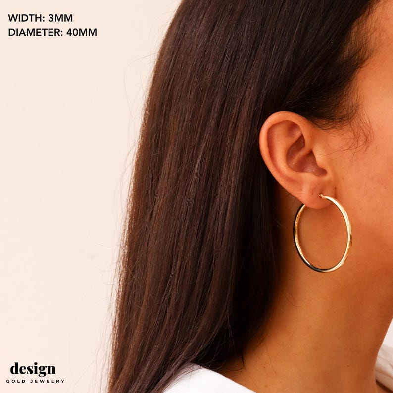 May include: Gold hoop earrings with a 40mm diameter and 3mm width. The earrings are shown on a person's ear.