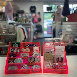 Puede incluir: Dos vitrinas de acrílico rosa llenas de parches bordados coloridos y joyas de cuentas. Los parches presentan varios diseños, incluyendo un sombrero de vaquero, un bronco y un lazo. Las joyas incluyen pulseras y collares de cuentas.