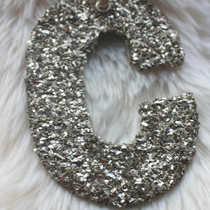 May include: A silver glitter-covered letter "C" ornament with a jute string for hanging. The letter is on a fluffy white background. This decorative item is suitable for holiday or home decor.