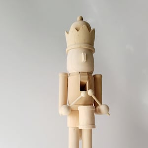 May include: A wooden nutcracker in the shape of a drummer, standing on a rectangular base. The nutcracker is unpainted, with a crown and drum. The drum is positioned at the waist, and drumsticks are held in each hand. The figure is approximately 12 inches tall.