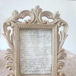 May include: A gold-colored ornate picture frame with a decorative design. The frame is sitting on a light brown surface. The frame contains a piece of paper with text in French. The text reads: "emplacer par danwief ue vous ferez vousnes Astringentellson  ementhet phiverelelove Comme lotion deux ou t le vinaigre d'alcão) et auta Mare de benjom dans un louce. Assouplissante et lenitive sion de camomille, suřeäu laurée mélangés en pers Infusionde racine de grin He laitue, infusion derfe son. vous v PulvérisationsSio pulvérisateur, v bierde Spratiquer Sur petit ez-1 les pulvérisations ledes av de énöfllent, eau d'oige par décoction de 1 eatu VOS N'artender à demein"