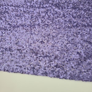 Puede incluir: Un primer plano de una tela de lentejuelas de color morado. Las lentejuelas son pequeñas y redondas, y están dispuestas en un patrón aleatorio. La tela es suave y brillante.