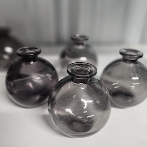 May include: Four small, round, gray glass vases with a smooth, shiny finish. The vases are arranged in a semi-circle on a white surface.
