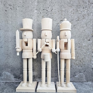 May include: Three unfinished wooden nutcracker figurines. The nutcracker on the left is holding a sword, the middle nutcracker is holding a drum, and the nutcracker on the right is holding a staff.