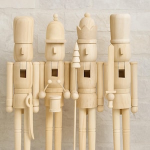 May include: Four wooden nutcrackers in a row, each with a unique hat and accessory. The nutcrackers are unpainted, showcasing the natural wood grain. One holds a sword, another a drum, and another a spear. They stand on individual wooden bases.