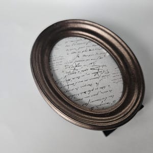 May include: An oval-shaped picture frame with a bronze finish. The frame has a decorative, raised edge and is standing on a black stand. The frame is empty, but the glass is visible and reflects the light.