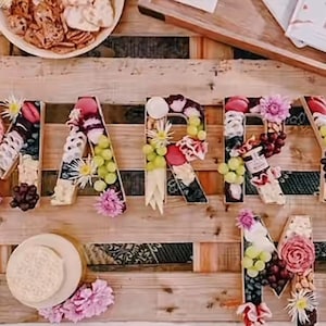 May include: Wooden letters spelling "MARRY ME" are decorated with cheese, grapes, flowers, and other charcuterie items. A wooden pallet background supports the letters and a plate of crackers. The letters are arranged for a wedding or engagement celebration.